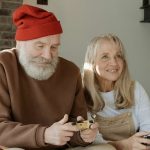 An old man with a red beanie sits on a sofa with an older blonde woman playing video games together.